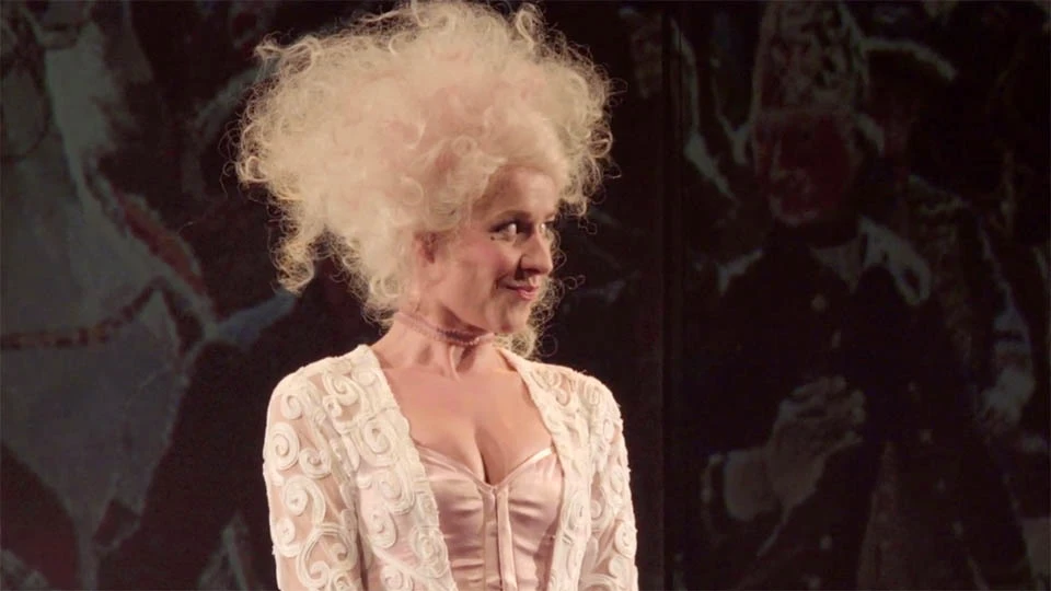 Eine Frau, die ein weißes, verziertes Kleid und eine dramatisch gepuderte weiße Perücke trägt, steht vor einem dunklen Hintergrund und blickt zur Seite.