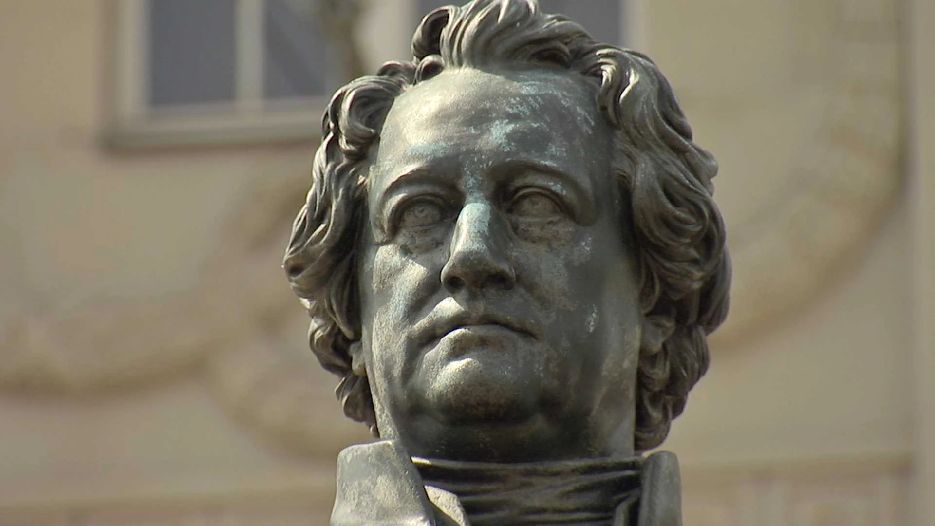 Nahaufnahme einer Bronzestatue, die den Kopf und die oberen Schultern eines Mannes mit gewelltem Haar darstellt, vor einem unscharfen Gebäudehintergrund.