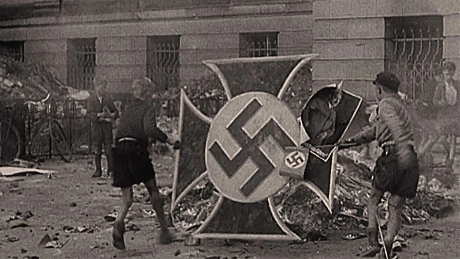 Drei Kinder zerschlagen ein großes Nazi-Symbol mit Stöcken auf einer Straße in der Stadt, mit Trümmern und einem Gebäude im Hintergrund.