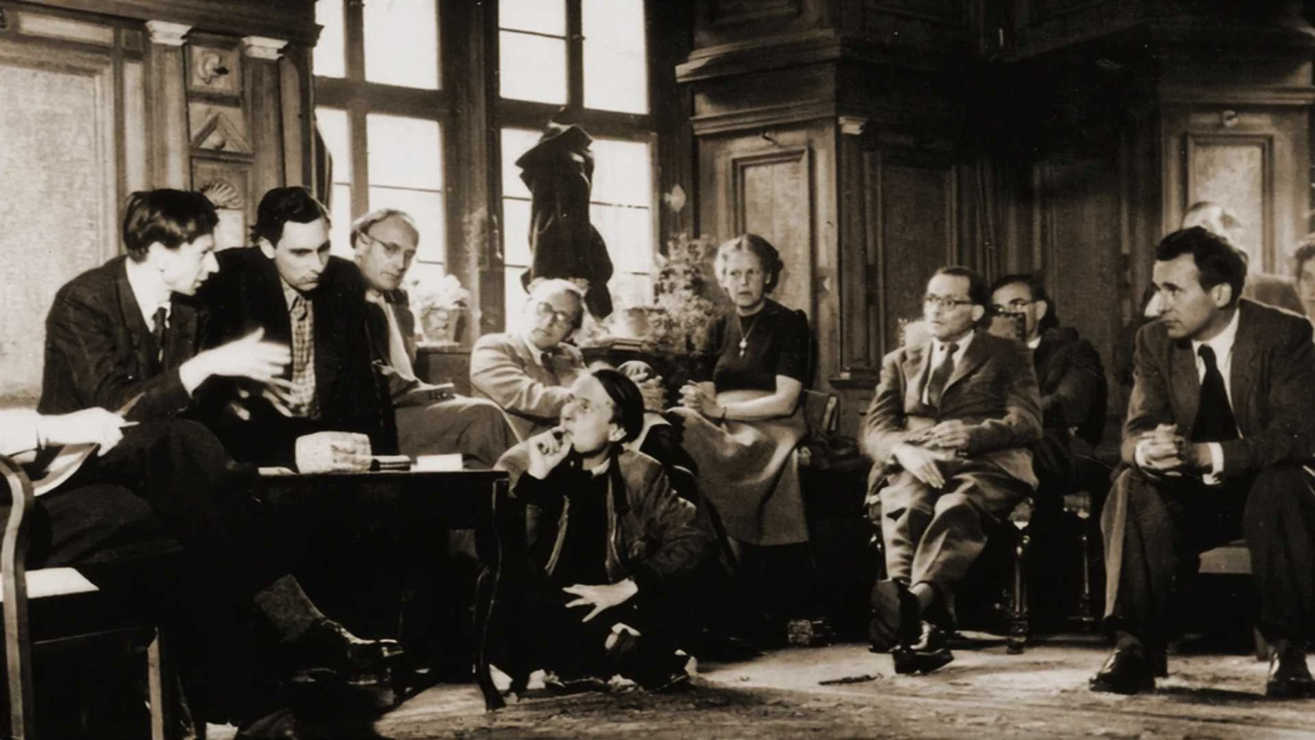 Eine Gruppe von Menschen sitzt in einem holzgetäfelten Raum mit großen Fenstern und unterhält sich; einige sitzen auf Stühlen, andere auf dem Boden, in einer lockeren, aufmerksamen Atmosphäre.