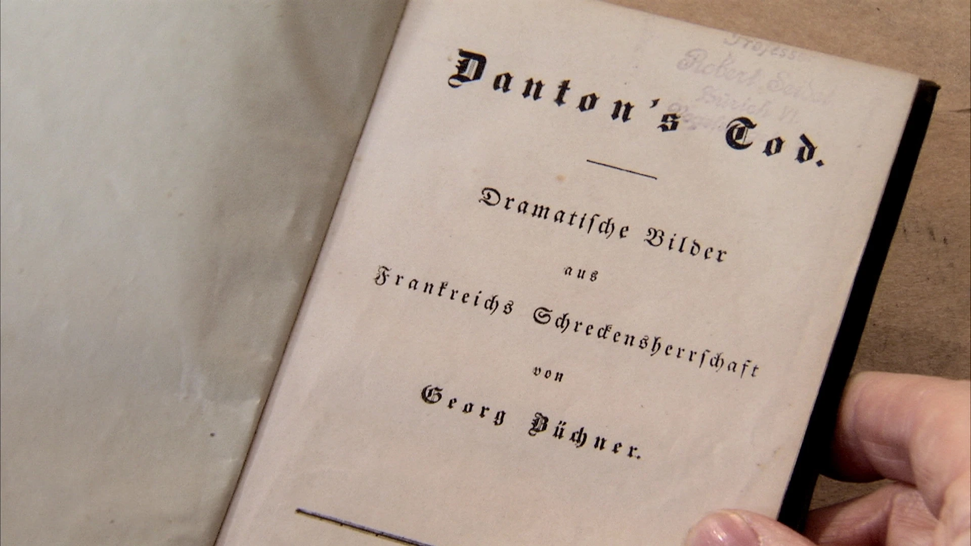Eine Hand hält ein aufgeschlagenes Buch mit dem Titel Dantons Tod. Dramatische Bilder aus Frankreichs Schreckensherrschaft von Georg Büchner.