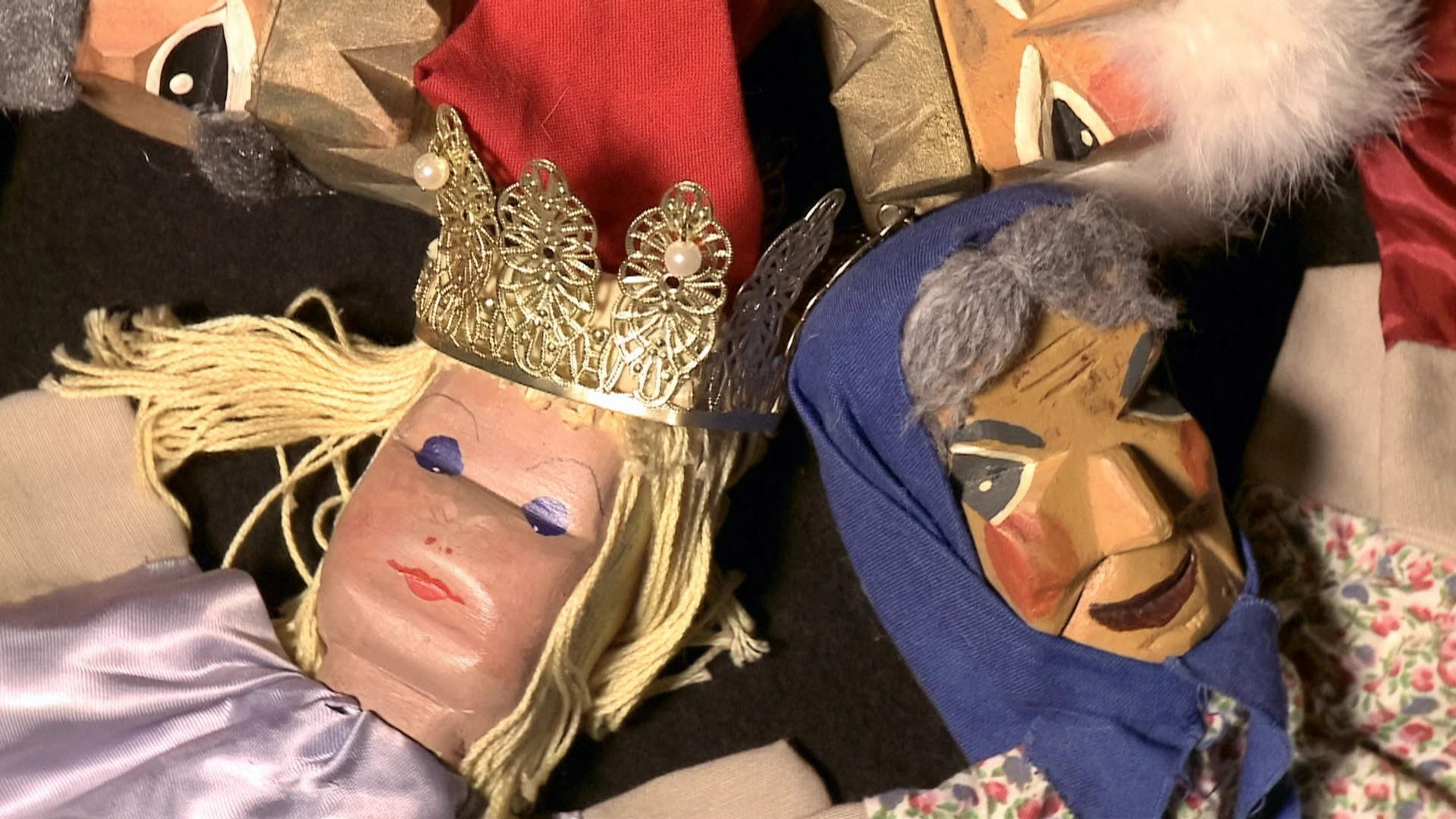 Nahaufnahme von zwei Holzpuppen; eine trägt eine silberne Krone und einen roten Hut, die andere ein blaues Kopftuch. Beide haben bemalte Gesichter und liegen auf einer dunklen Fläche.