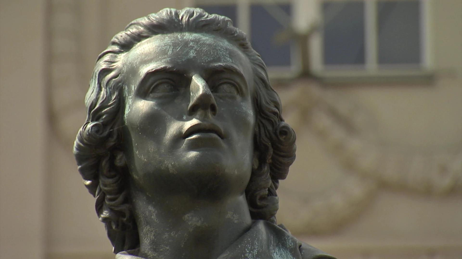 Nahaufnahme einer Bronzestatue, die eine Person mit gewelltem Haar darstellt, die leicht nach oben schaut; unscharfer architektonischer Hintergrund.