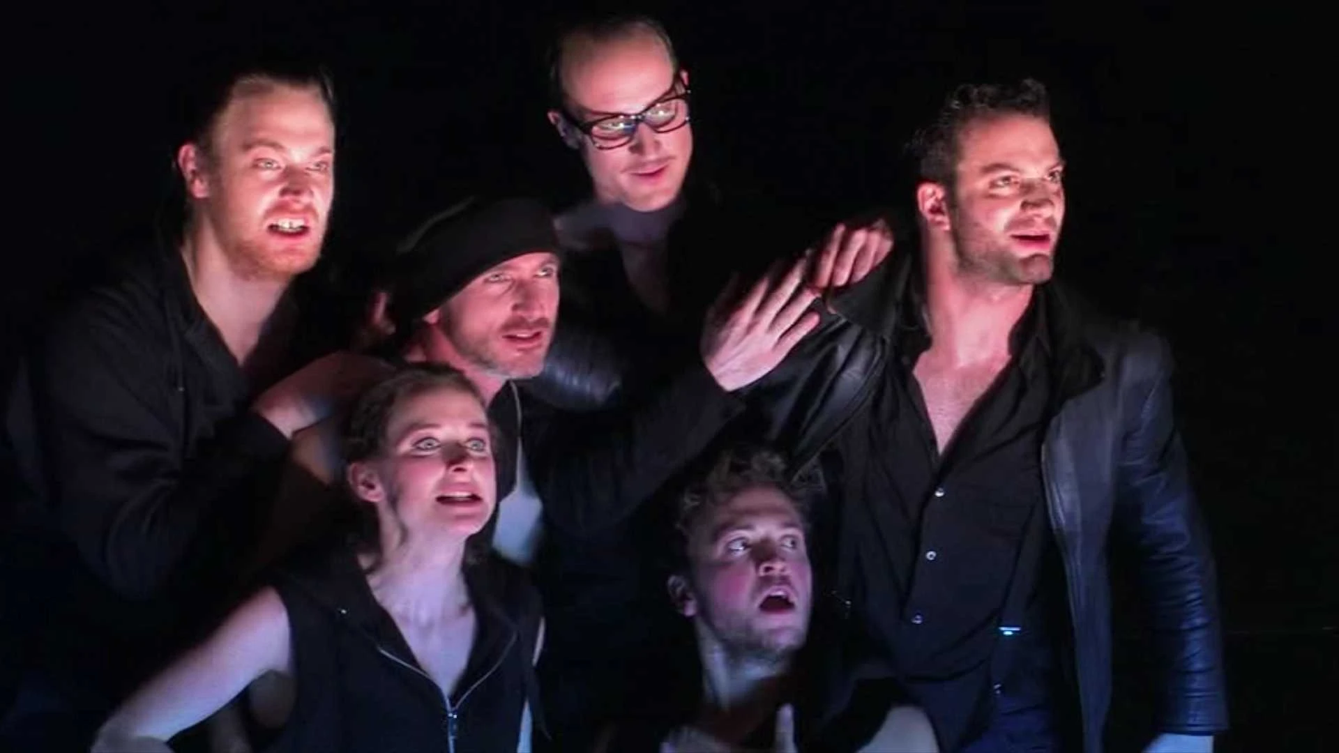 Sechs dunkel gekleidete Schauspieler stehen dicht beieinander auf der Bühne und schauen bei dramatischer Beleuchtung intensiv in die gleiche Richtung.