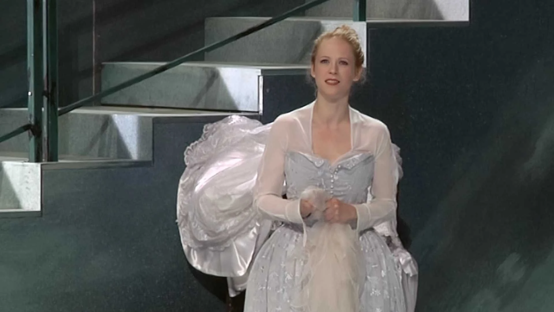 Eine Frau in einem hellblauen Ballkleid steht vor einer Treppe, die Hände vor sich verschränkt.