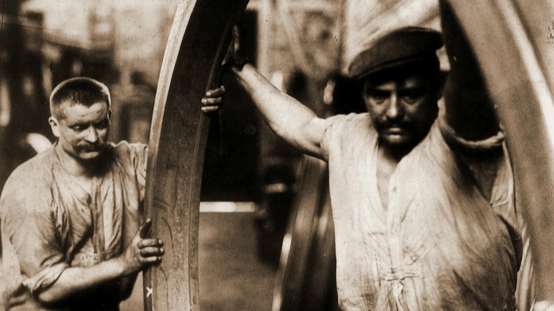 Zwei Männer in Arbeitskleidung hantieren mit einem großen Metallring in einer industriellen Umgebung, wahrscheinlich einer Fabrik oder Werkstatt.