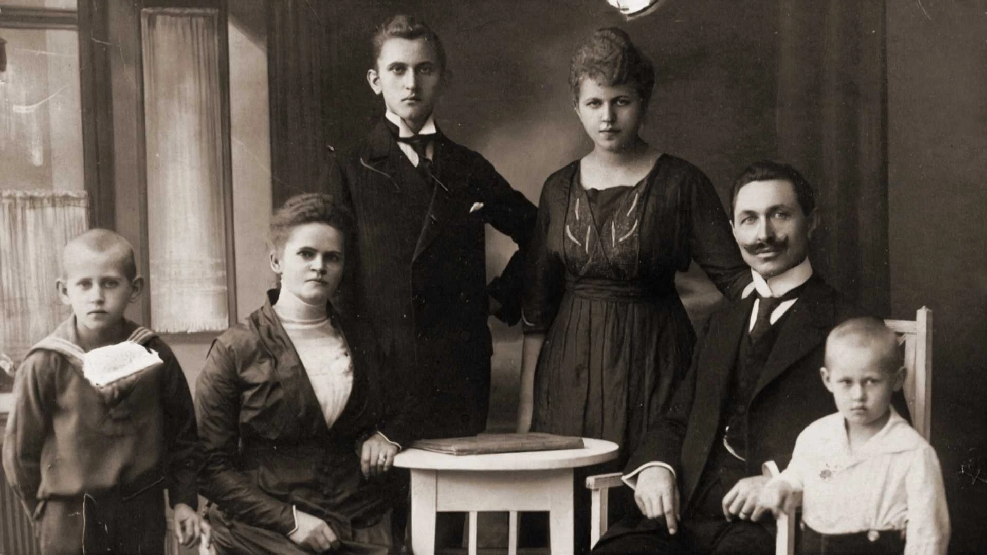 Ein altes Schwarz-Weiß-Foto von sechs Personen - zwei erwachsene Männer, zwei erwachsene Frauen und zwei kleine Jungen -, die in einem Haus für ein formelles Familienporträt posieren.