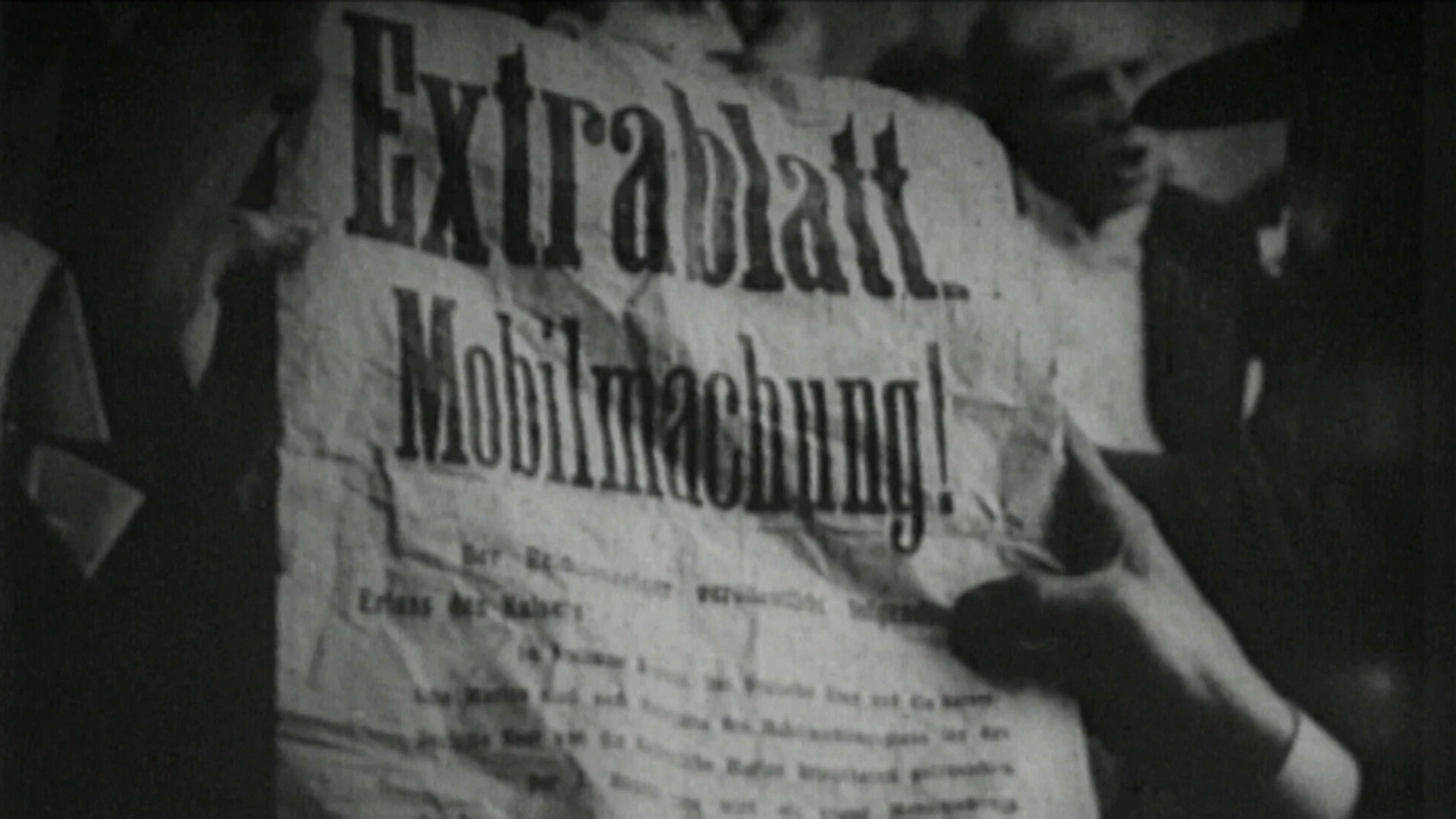 Eine Person hält eine zerknitterte Zeitung mit der deutschen Schlagzeile Extrablatt Mobilmachung! in einer schwarz-weißen Szene mit mehreren Personen im Hintergrund.