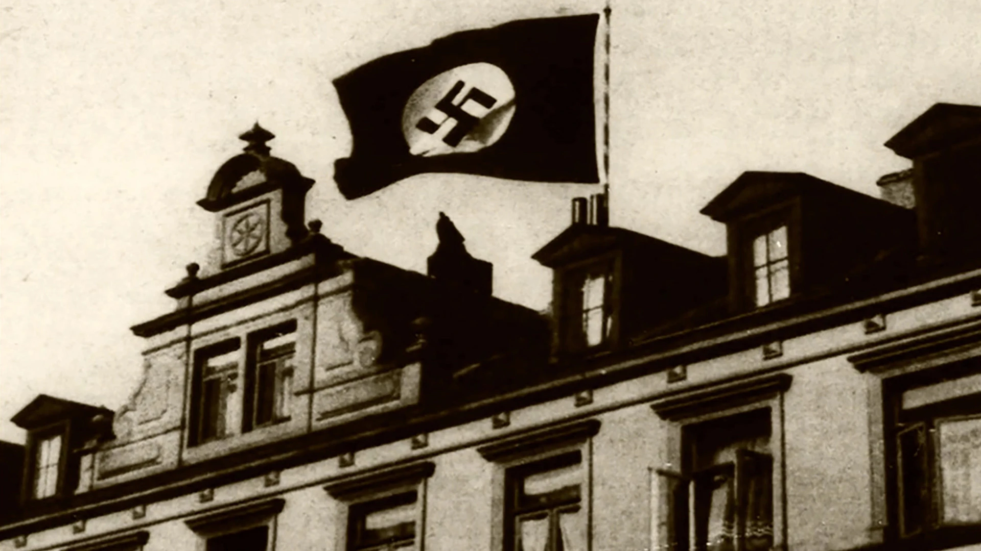 Ein Gebäude mit einer Hakenkreuzfahne, die mit Nazi-Deutschland assoziiert wird, weht von seinem Dach. Das Foto scheint historisch zu sein, in schwarz-weiß.
