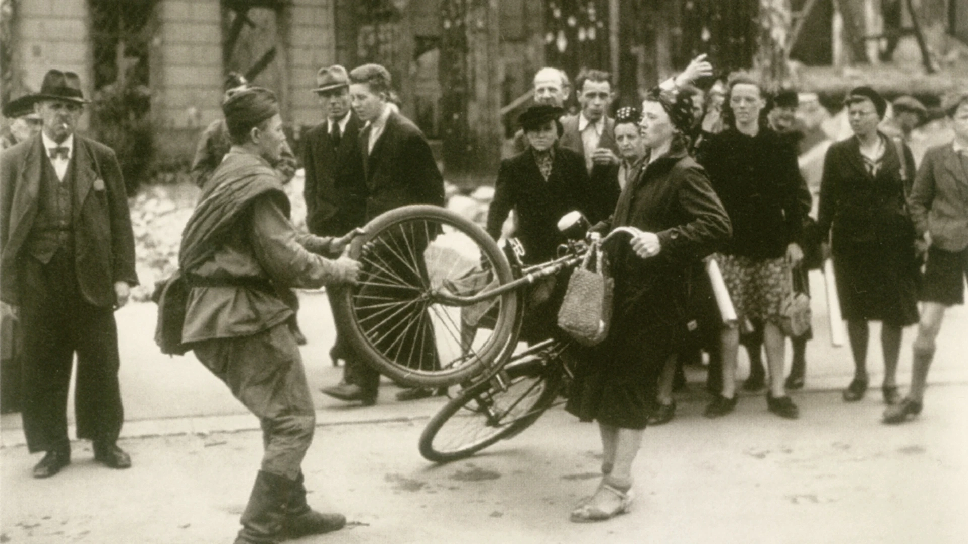 Ein Soldat nimmt einer Frau gewaltsam ein Fahrrad ab, während eine Gruppe von Schaulustigen auf einer Straße in der Stadt zusieht, mit beschädigten Gebäuden im Hintergrund.