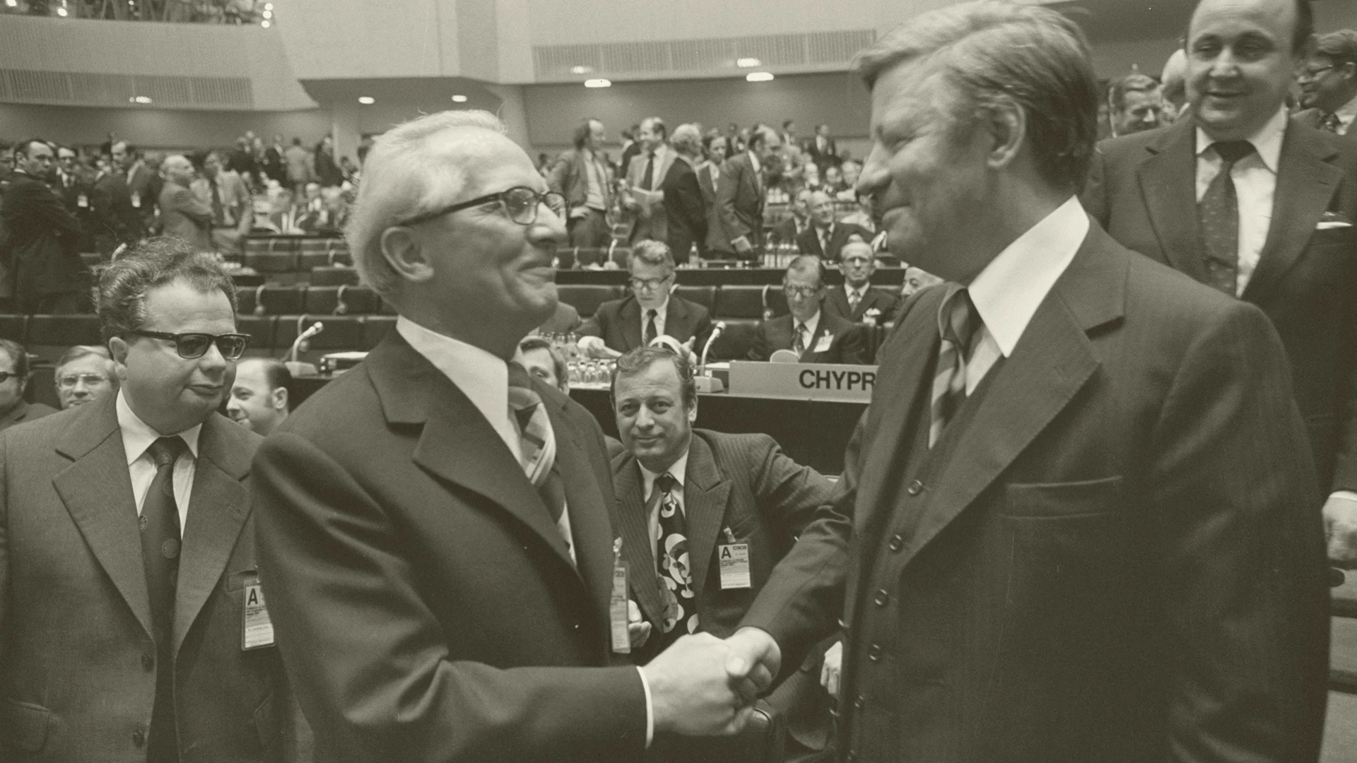 Zwei Männer in Anzügen schütteln sich die Hände und lächeln in einem überfüllten Konferenzsaal, in dem im Hintergrund Teilnehmer und reservierte Namensschilder zu sehen sind.