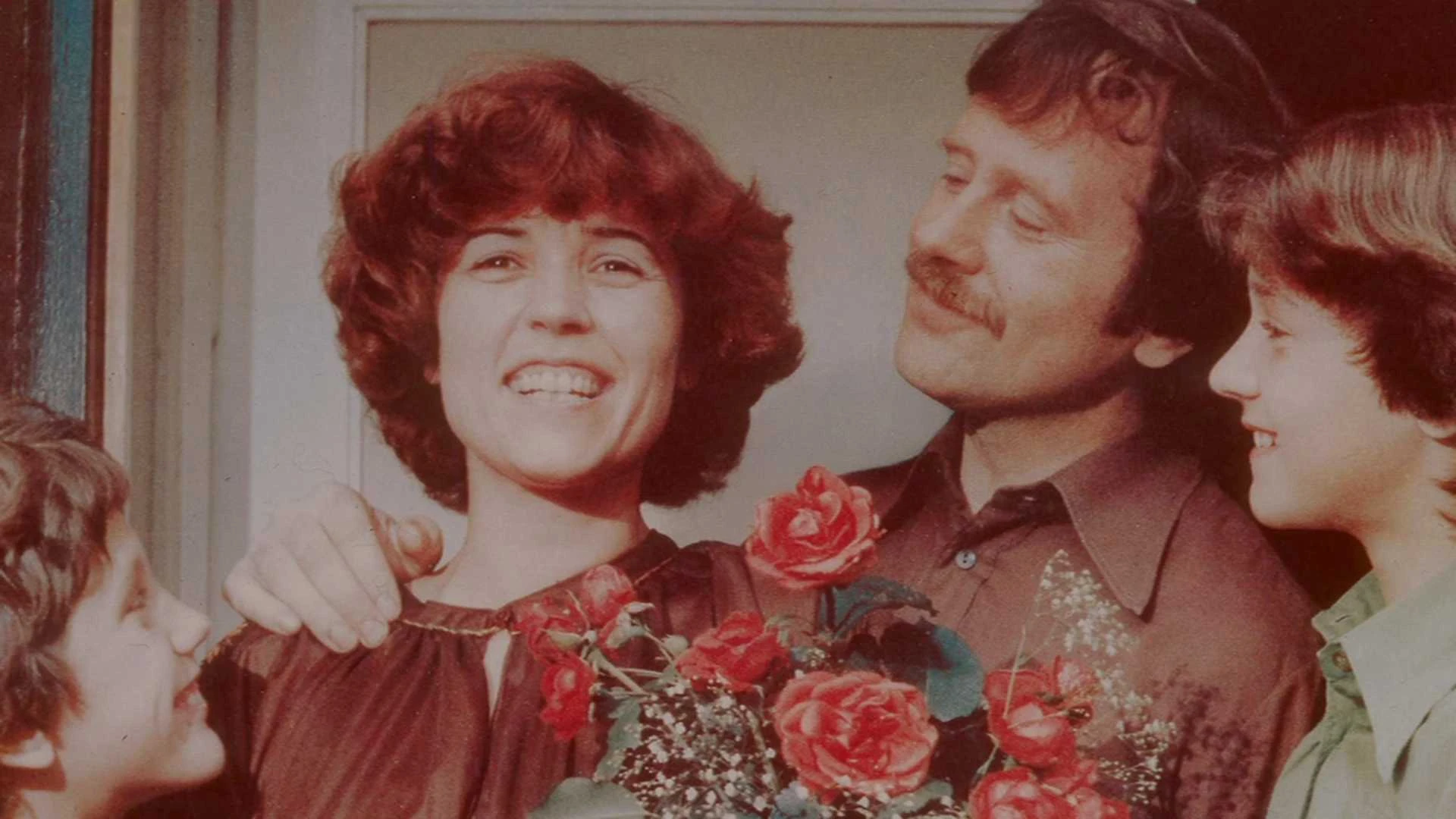 Eine Frau lächelt, während sie einen Strauß roter Rosen in der Hand hält, umgeben von einem Mann und zwei Kindern, die alle zusammen glücklich zu sein scheinen.