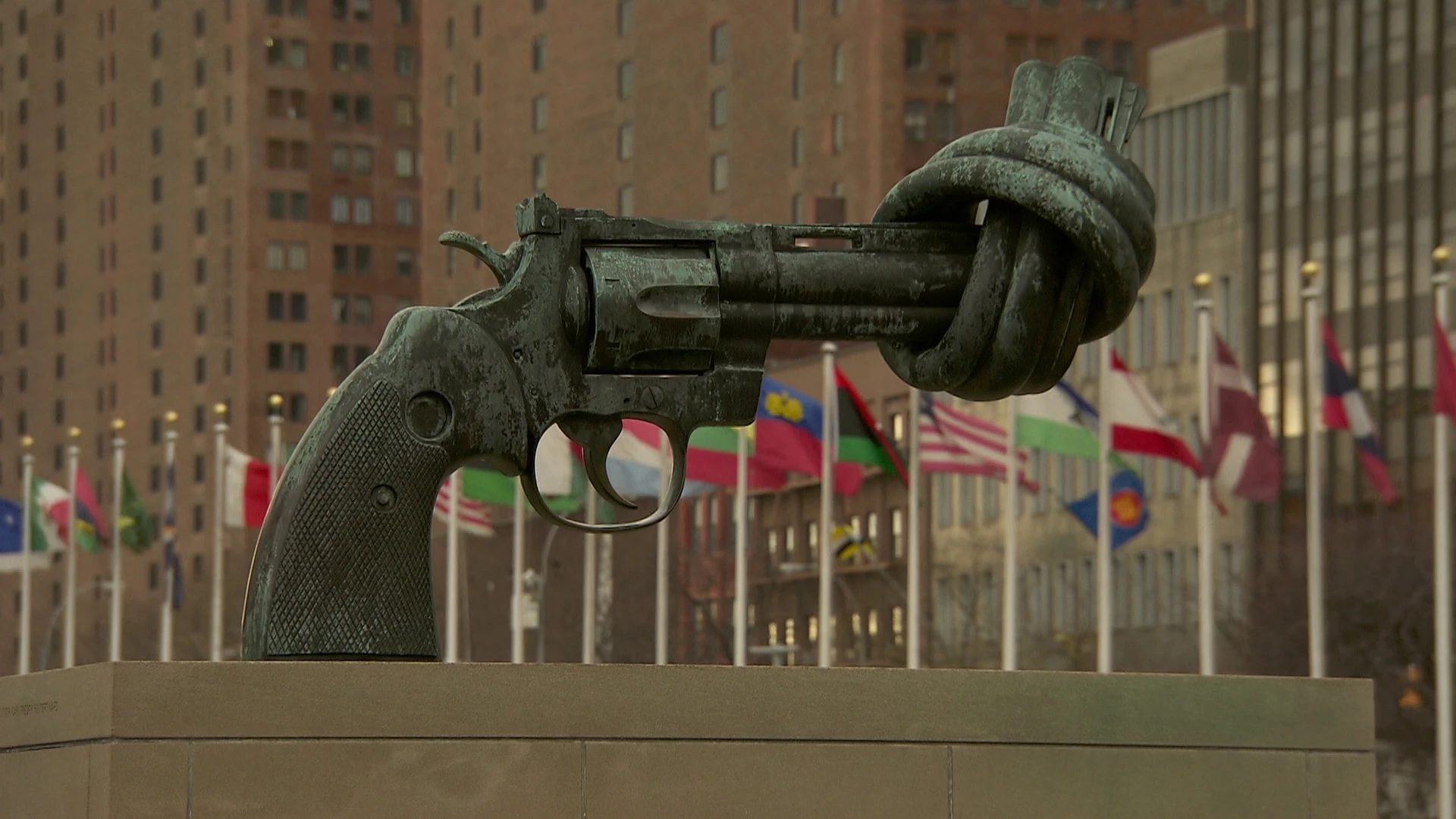 Eine Bronzeskulptur eines Revolvers mit verknotetem Lauf, die im Freien vor zahlreichen internationalen Flaggen und hohen Gebäuden aufgestellt ist.