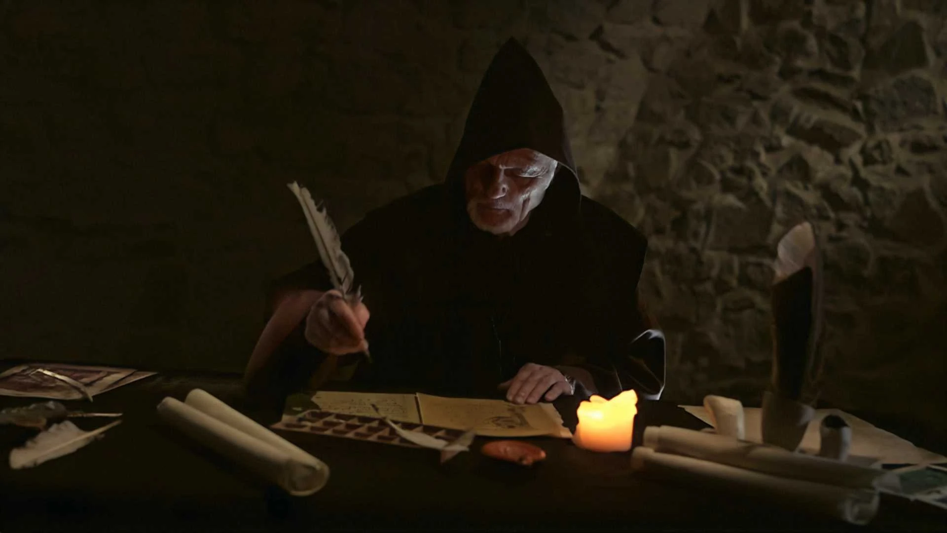 Eine Person in einem dunklen Kapuzenmantel schreibt bei Kerzenlicht mit einer Schreibfeder an einem Tisch mit Schriftrollen und Papieren vor einem Hintergrund aus Steinmauern.