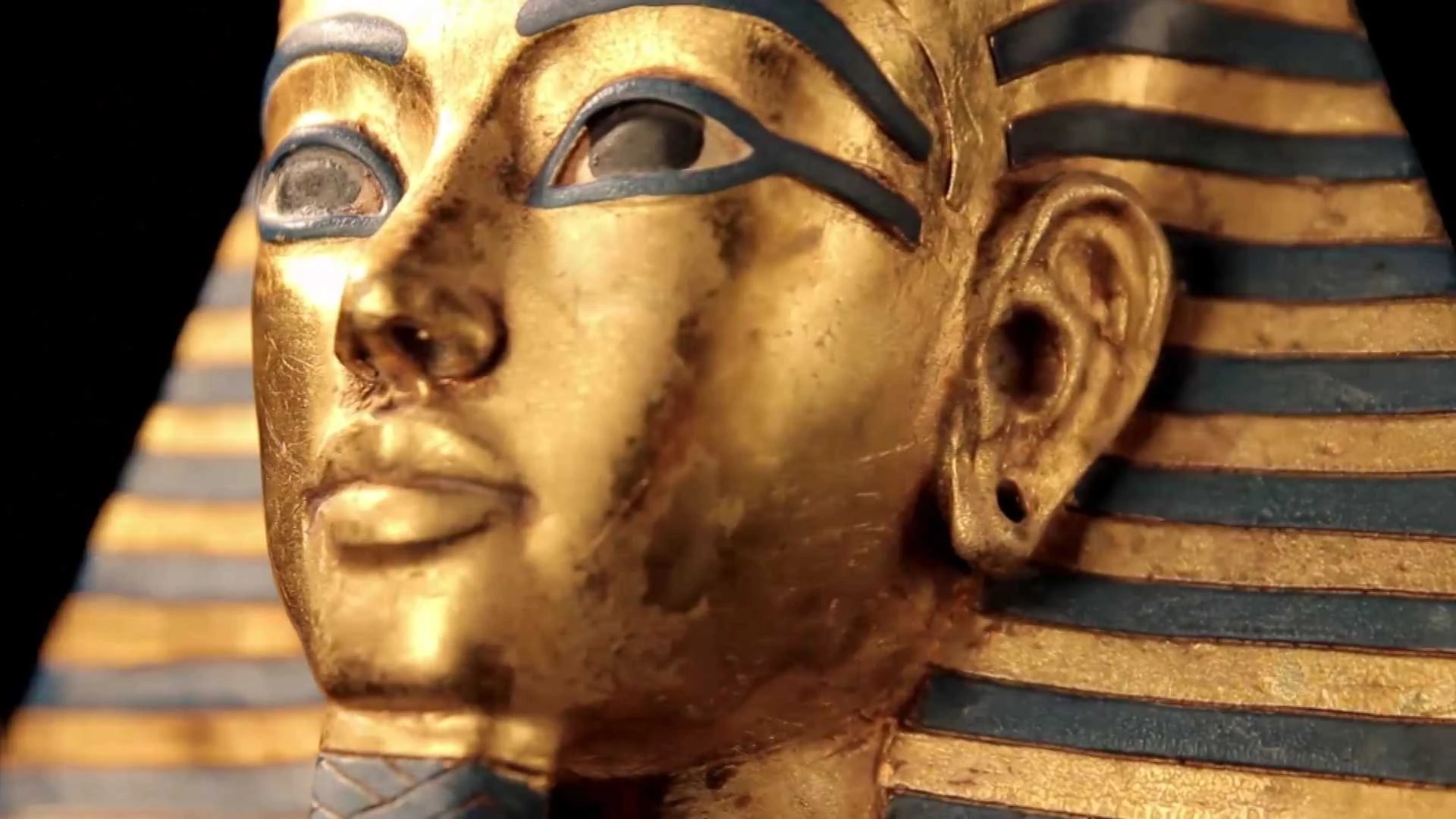 Nahaufnahme der goldenen Maske eines altägyptischen Pharaos mit blau-gold gestreiftem Kopfschmuck und detaillierten Gesichtszügen.