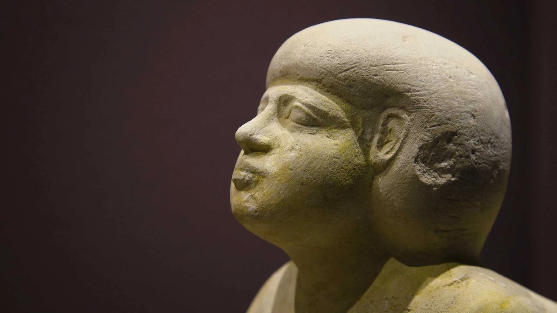 Nahaufnahme einer antiken Steinstatue, die eine Person mit einer runden Frisur, markanten Gesichtszügen und einem ruhigen Gesichtsausdruck vor einem dunklen Hintergrund darstellt.