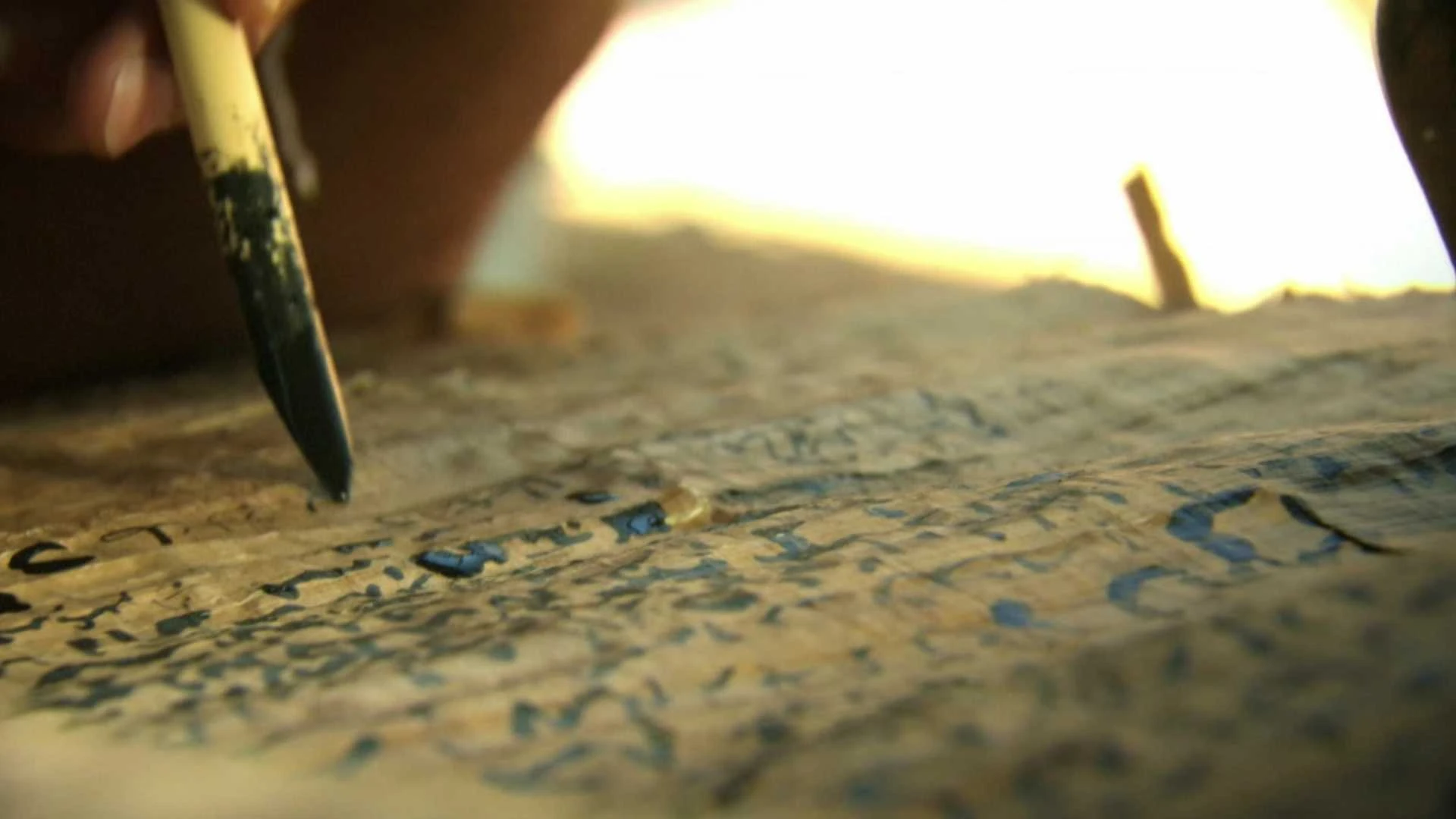Nahaufnahme einer Hand, die mit einem Pinsel oder Stift blaue und schwarze Symbole auf eine alte Papyrusrolle schreibt oder malt.