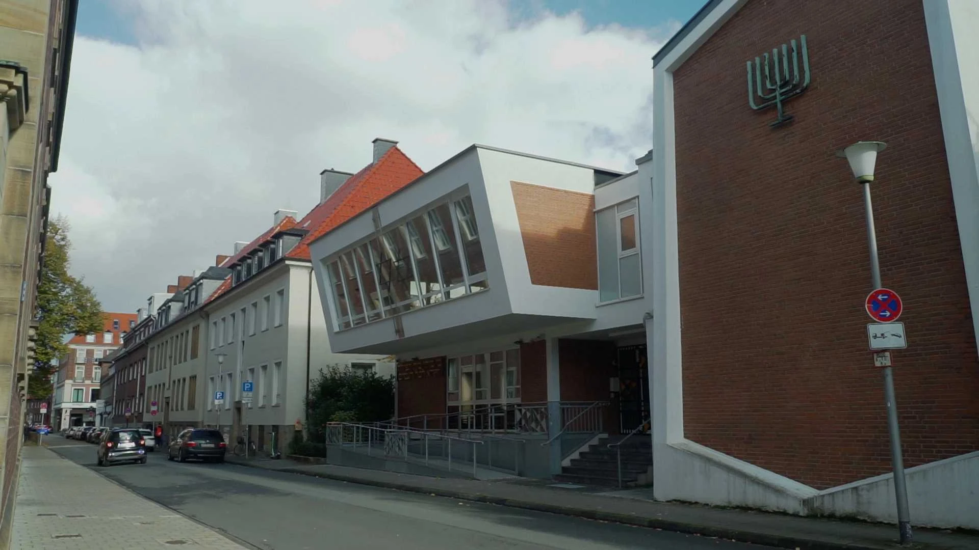 Eine moderne Synagoge mit einem markanten Überbau und einem Menora-Symbol an der Außenwand steht an einer schmalen Straße, die von parkenden Autos und Wohnhäusern gesäumt ist.