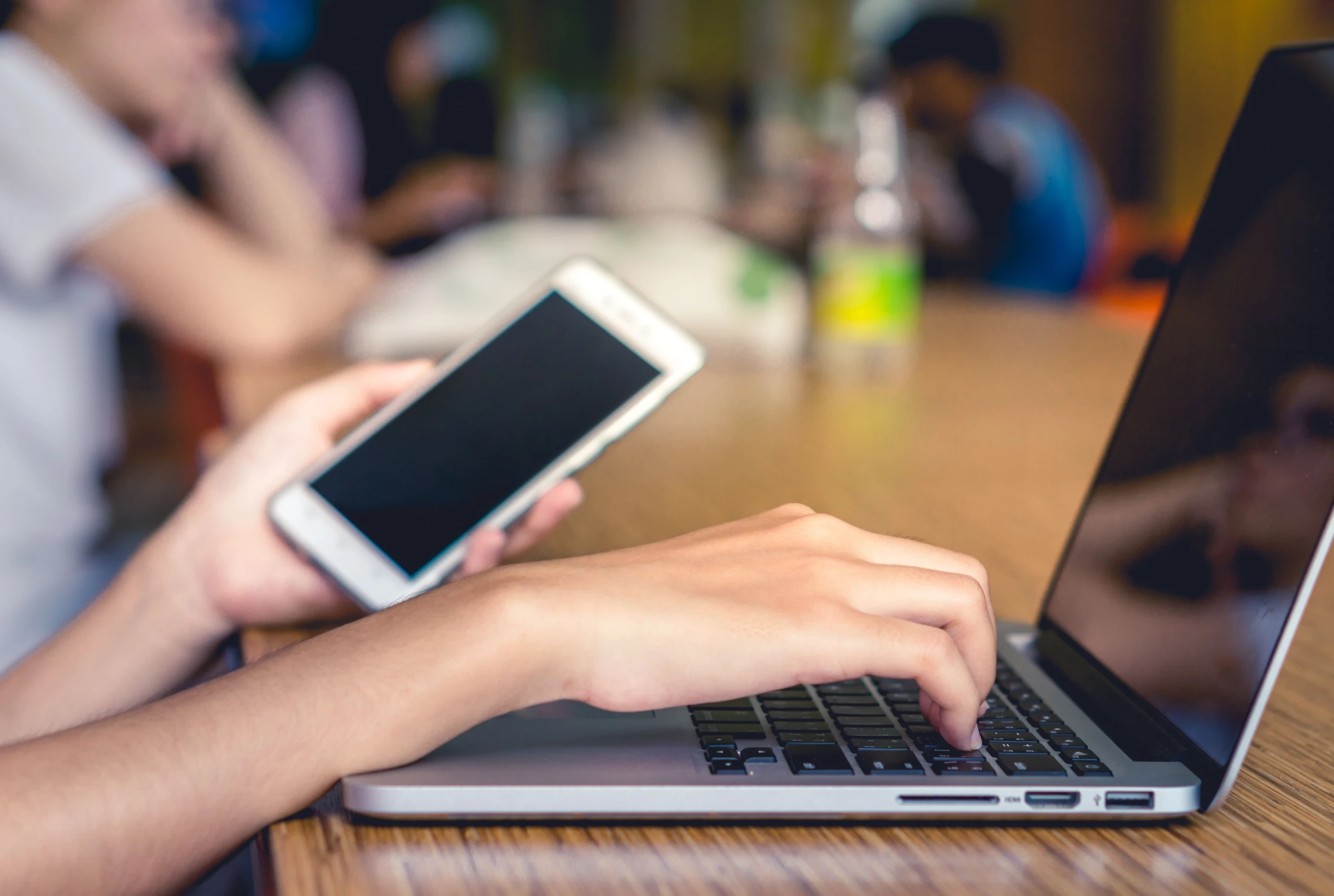 Eine Person sitzt an einem Tisch und bedient mit der einen Hand einen Laptop, während sie in der anderen Hand ein Smartphone hält, mit unscharfen Personen und Objekten im Hintergrund.